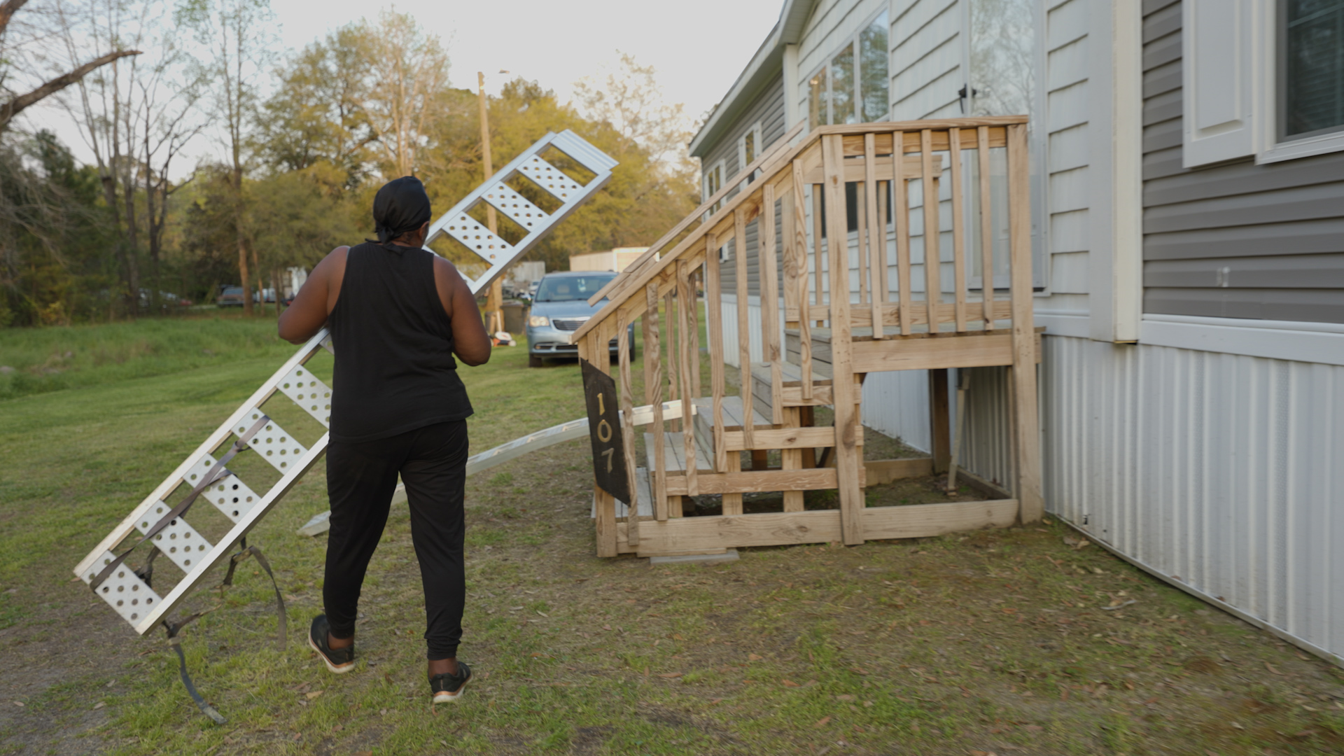 Dolores carrying the aluminum ATV ramps toward the steps, back to camera